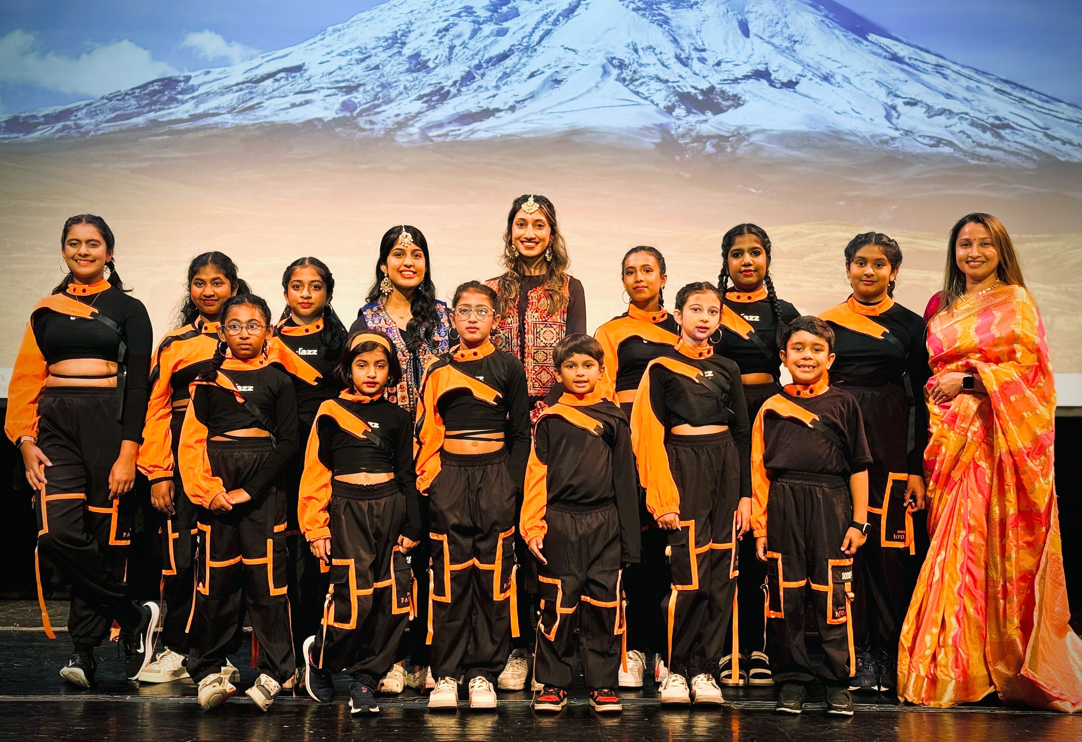 Garima Dance Academy students and instructor in black-and-orange costumes, mountain stage backdrop